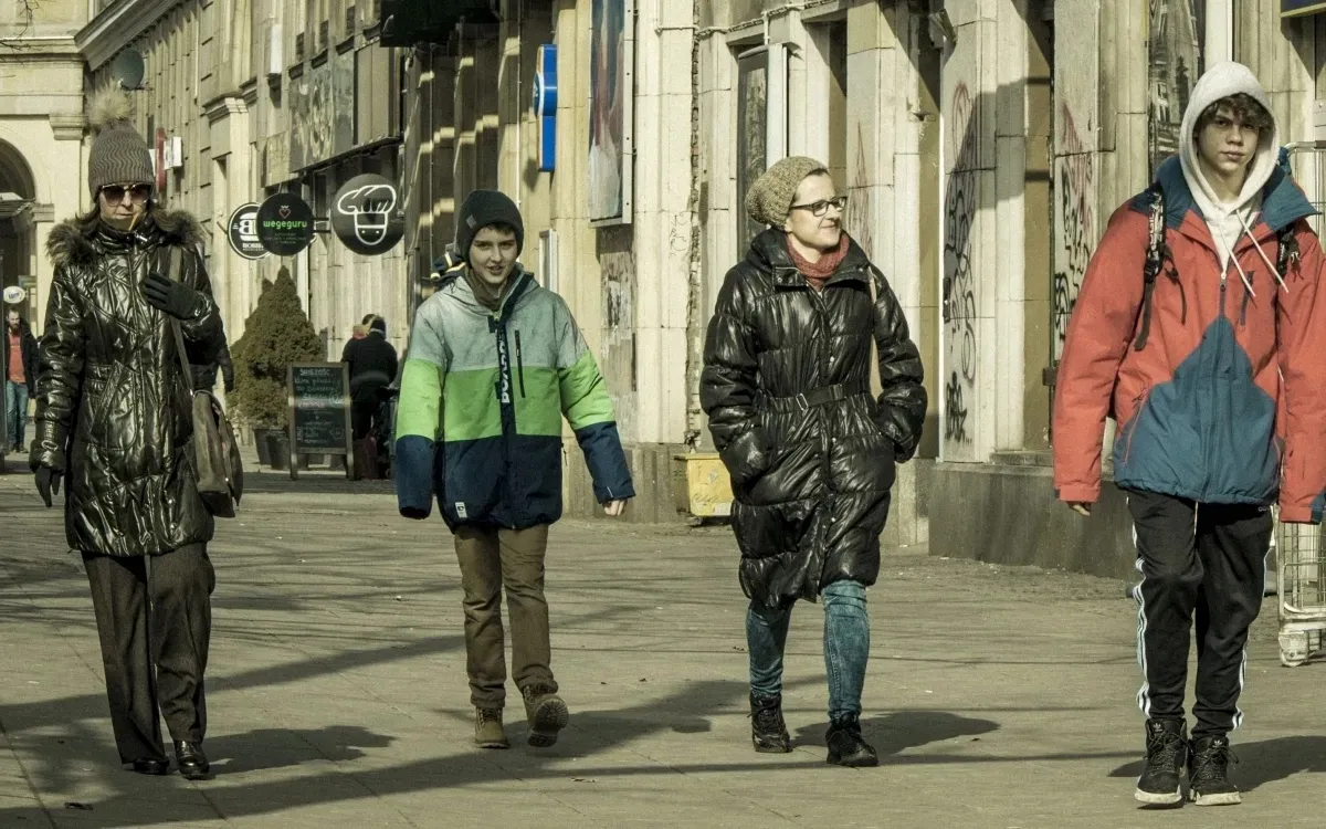 Four pedestrians walking along a sunlit street in winter coats